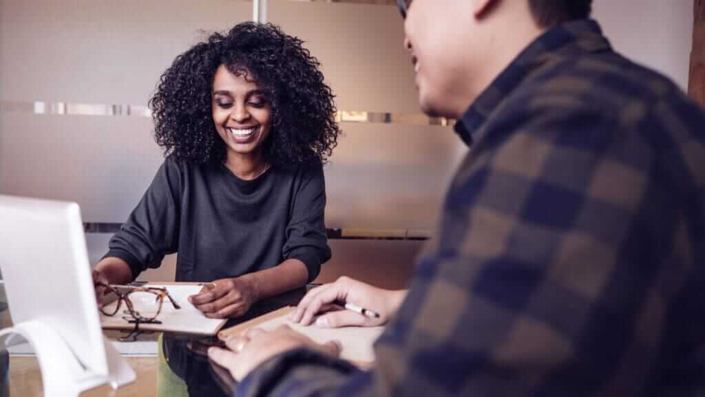 Two people are seated at a table in an office setting. One person with curly hair is smiling and holding a pencil. The other, whose back is partially facing the camera, wears glasses and a plaid shirt. A computer monitor is visible on the table, hinting at their focus on small business sustainability strategies.