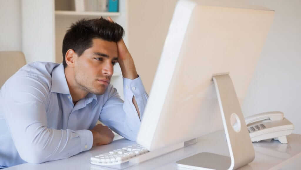 A man in a blue shirt is sitting at a desk, leaning his head on one hand, staring intently at a large computer monitor. Despite his frustration or fatigue, his focus and determination exhibit clear leader characteristics. There is a keyboard, mouse, and phone on the desk in the office setting.