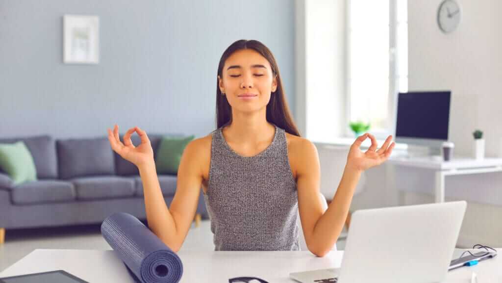 A woman sits at a desk in a home office with her eyes closed, hands in a meditative pose, likely reflecting on her small business sustainability goals. A yoga mat lies rolled up beside her. Behind her, a living room with a gray sofa and several cushions is visible. A clock hangs on the white wall in the background.