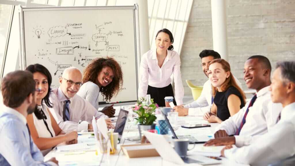 A diverse group of professionals is sitting and standing around a conference table, with laptops, papers, and coffee cups, engaged in a discussion. A woman stands at the head of the table, smiling. Behind her is a whiteboard with diagrams and notes focused on small business sustainability.