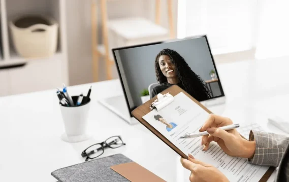 A recruiter reviews a resume on a clipboard while conducting a video interview with a woman shown on a laptop screen.