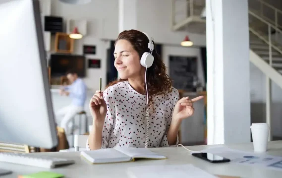 Woman wearing headphones, smiling and dancing while working at her desk, enjoying music during work.