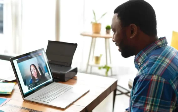 A man in a plaid shirt smiles while attending a video call on his laptop at a home office desk.