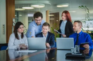 A group of professionals collaborate around a conference table with laptops in a modern office setting, representing teamwork and productivity.