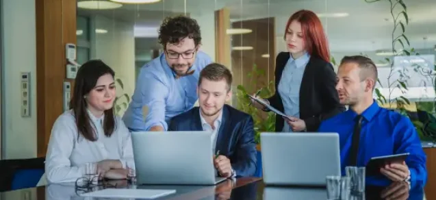 A group of professionals collaborate around a conference table with laptops in a modern office setting, representing teamwork and productivity.