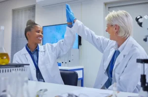 Two scientists in lab coats and gloves high-five in a modern laboratory, celebrating a successful experiment amid scientific equipment and glassware.