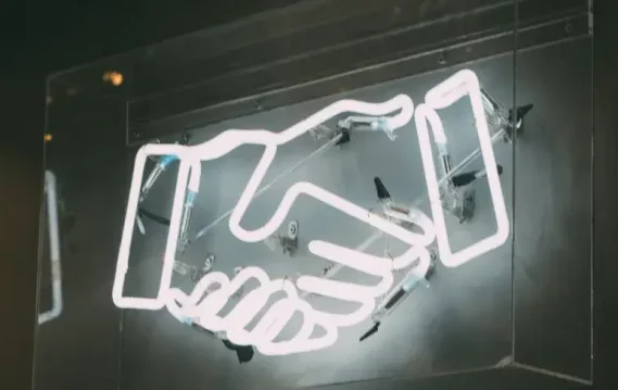 A glowing neon sign of two hands shaking, symbolizing agreement and partnership, against a dark background.