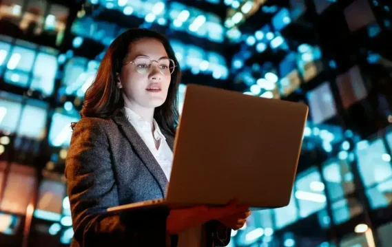 Businesswoman holding laptop at night in front of illuminated city building, suggesting technology and modern work culture.