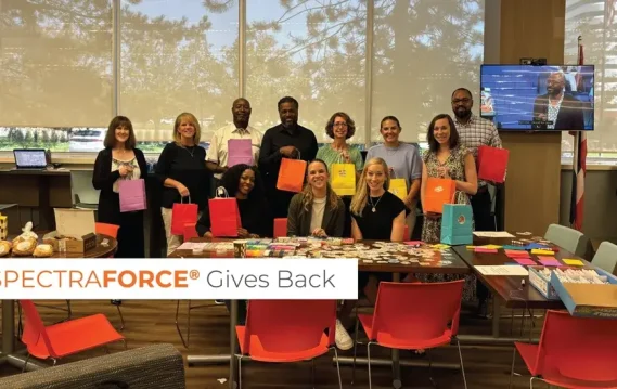 A group of employees holds colorful gift bags in a bright office setting, with decorated tables and baked goods, symbolizing the charitable efforts of SPECTRAFORCE in support of Grace Centers of Hope.