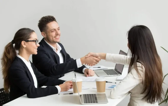 A woman in a white blazer shakes hands with a male interviewer while a female interviewer looks on, all smiling during a job interview.