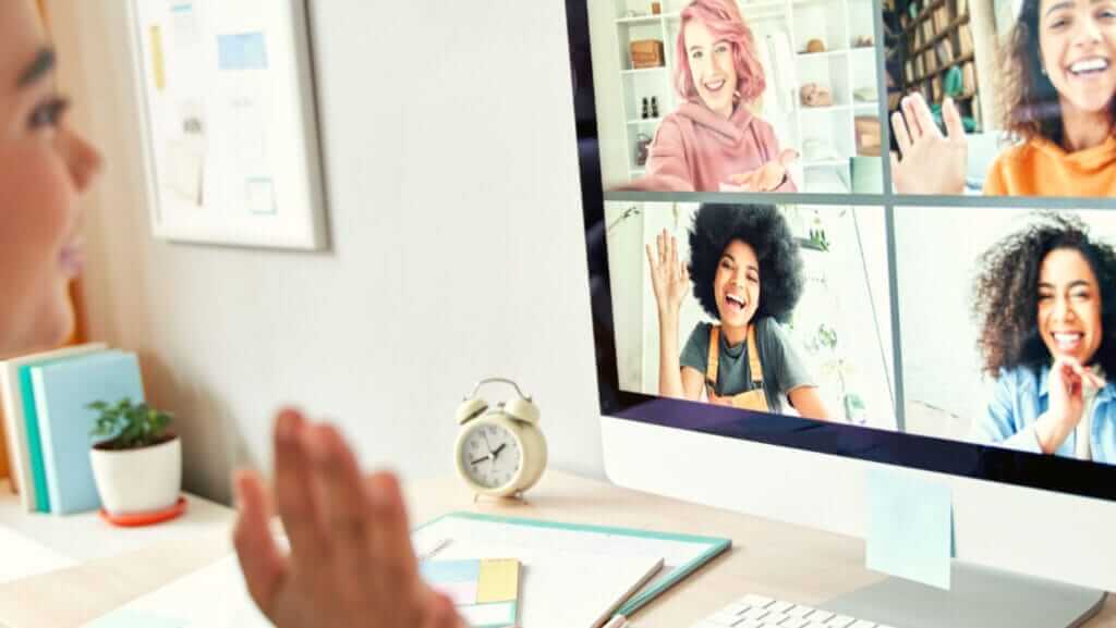 A person with leader characteristics participates in a video call on a computer, waving at four smiling individuals displayed on the screen. The scene includes a desk with a notebook, pens, and an alarm clock. Shelves and plants add to the cozy background.