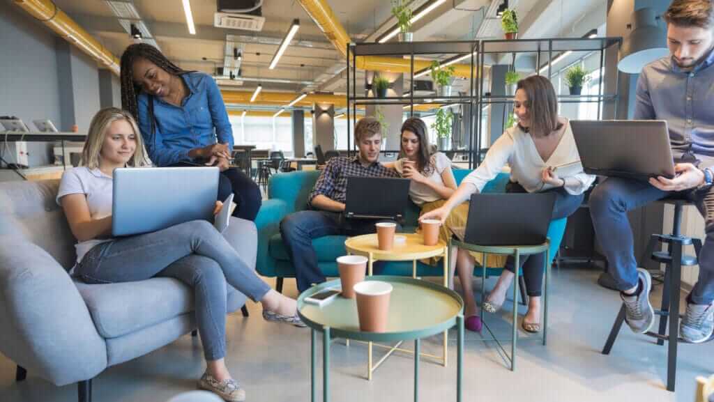 A group of six young adults, showcasing their talent in 2022, work on laptops in a modern, collaborative office space. They are seated on casual furniture, including armchairs and stools. The space features open shelves with plants and a combination of natural and industrial decor.